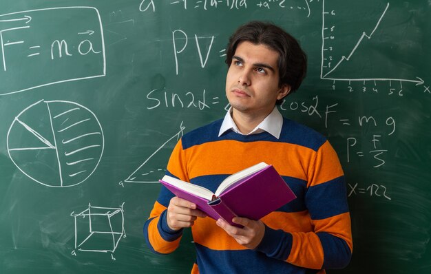 Thoughtful young caucasian geometry teacher standing in front of chalkboard in classroom holding book looking up