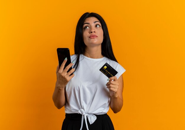 Thoughtful young caucasian girl holds phone and credit card looking at side 