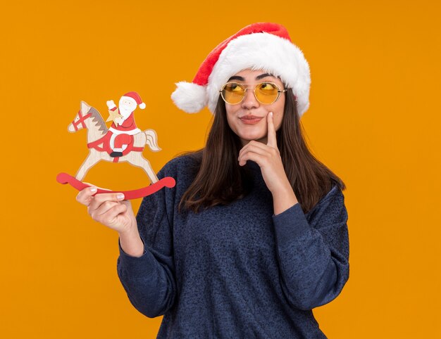 Thoughtful young caucasian girl in sun glasses with santa hat puts finger on chin and holds santa on rocking horse decoration looking up isolated on orange wall with copy space