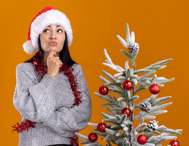 思いやりのある若い白人の女の子の首の周りにクリスマス帽子と見掛け倒しの花輪を身に着けているオレンジ色の背景で隔離の見上げるあごに触れる装飾されたクリスマスツリーの近くに立っている