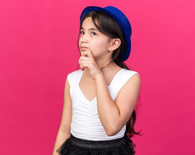 thoughtful young caucasian girl with blue party hat holding chin and looking up isolated on pink wall with copy space