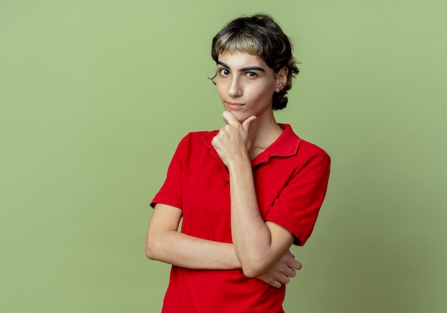 Free photo thoughtful young caucasian girl with pixie haircut putting hand on chin isolated on olive green background with copy space