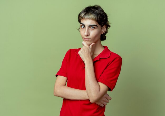 Thoughtful young caucasian girl with pixie haircut putting hand on chin isolated on olive green background with copy space