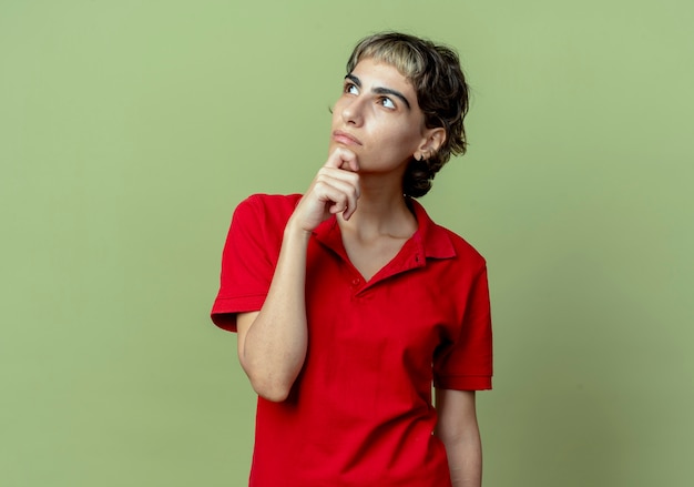 Free photo thoughtful young caucasian girl with pixie haircut putting hand on chin looking up isolated on olive green background with copy space