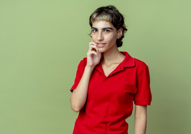 Thoughtful young caucasian girl with pixie haircut touching face isolated on olive green background with copy space