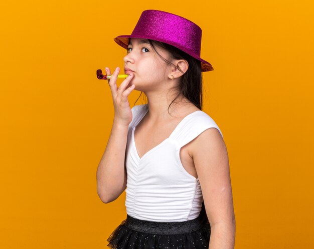 вдумчивая молодая кавказская девушка в фиолетовой шляпе смотрит вверх, держа партийный свисток, изолированную на оранжевой стене с копией пространства
