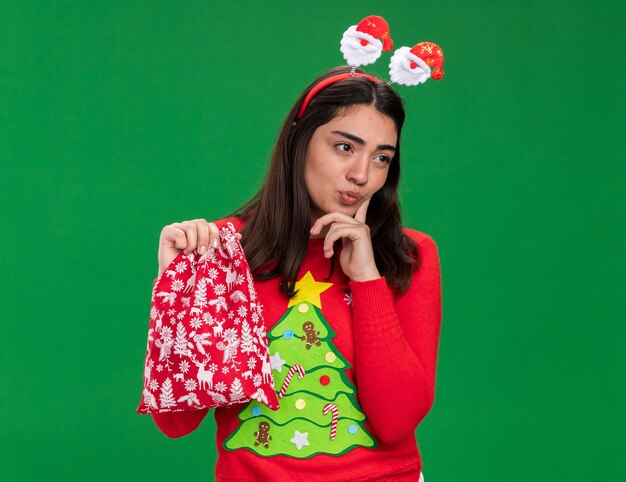 산타 머리띠와 사려 깊은 젊은 백인 여자는 턱에 손가락을 넣고 복사 공간이 녹색 배경에 고립 된 측면을보고 크리스마스 선물 가방을 보유
