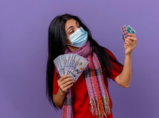 Thoughtful young caucasian ill girl wearing mask and scarf holding money and pack of capsules looking at capsules isolated on purple wall with copy space