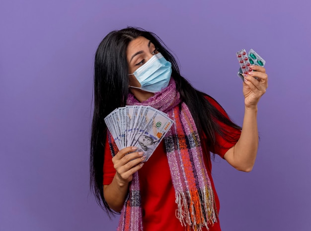 Thoughtful young caucasian ill girl wearing mask and scarf holding money and pack of capsules looking at capsules isolated on purple wall with copy space