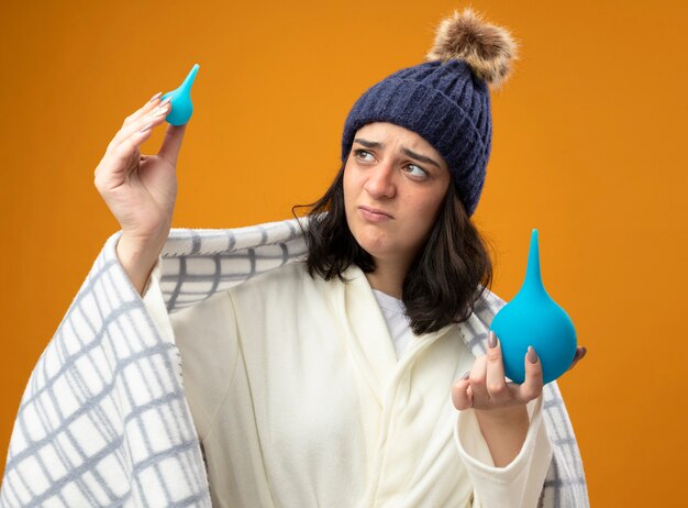 Thoughtful young caucasian ill girl wearing robe winter hat wrapped in plaid holding enemas raising up and looking at little one isolated on orange background