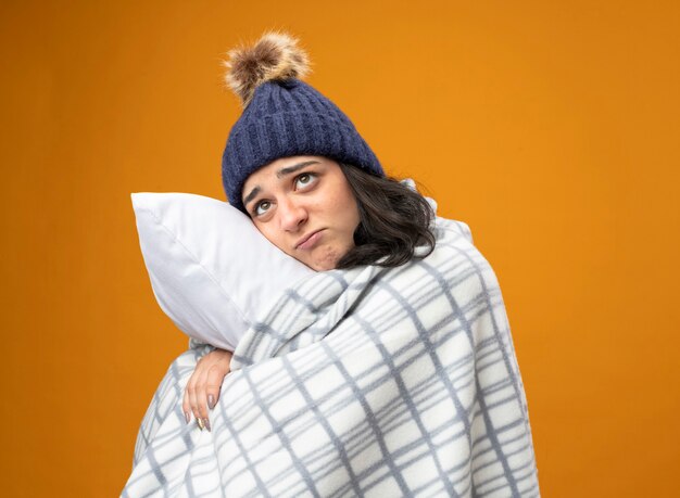 Thoughtful young caucasian ill girl wearing robe winter hat wrapped in plaid standing in profile view hugging pillow looking up isolated on orange background