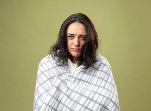 Thoughtful young caucasian ill girl wearing robe wrapped in plaid looking at camera grabbing plaid isolated on olive green background with copy space