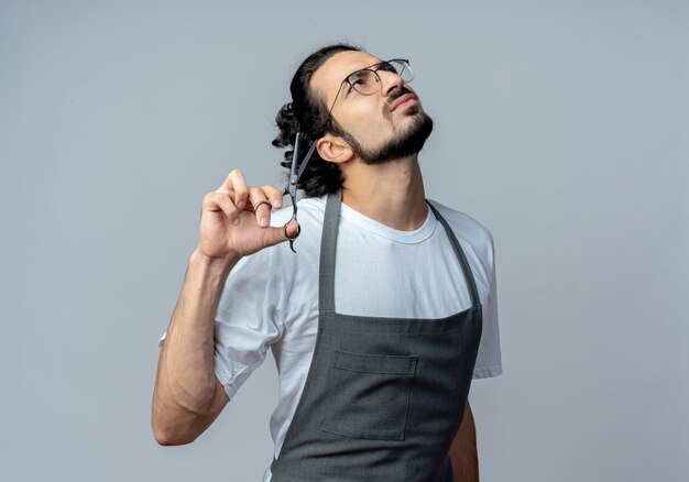 Thoughtful young caucasian male barber wearing glasses and wavy hair band in uniform holding scissors looking up isolated on white background with copy space