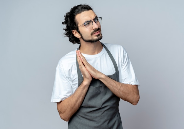 Thoughtful young caucasian male barber wearing glasses and wavy hair band in uniform looking at side and keeping hands together isolated on white background with copy space