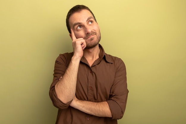 Thoughtful young caucasian man looking up pointing finger on temple isolated on olive green background with copy space