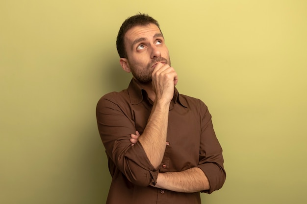 Thoughtful young caucasian man putting hand on chin looking up isolated on olive green background with copy space
