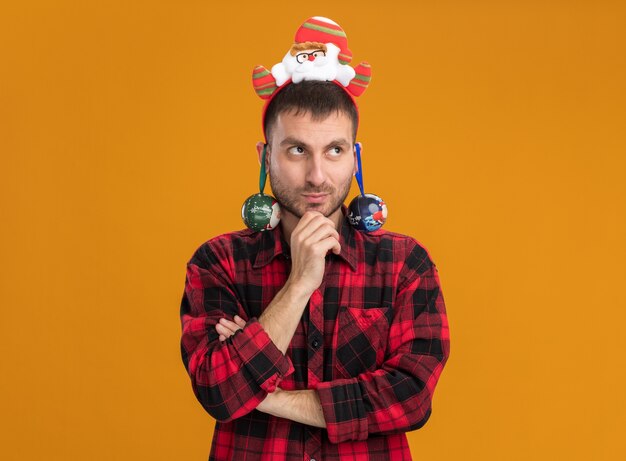 Thoughtful young caucasian man wearing santa claus headband touching chin looking at side with christmas baubles hanging from his ears isolated on orange background with copy space