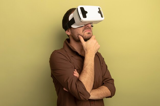 Thoughtful young caucasian man wearing vr headset putting hand on chin looking straight isolated on olive green background with copy space