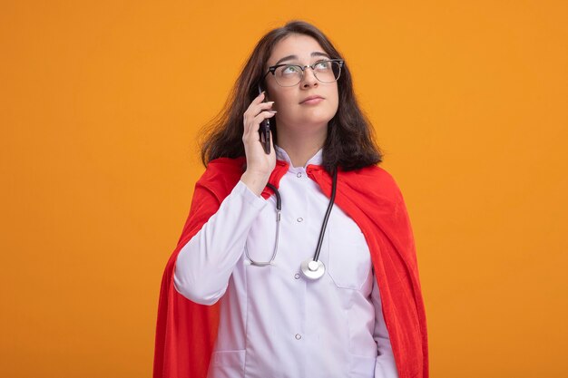 Thoughtful young caucasian superhero girl in red cape wearing doctor uniform and stethoscope with glasses talking on phone looking up 