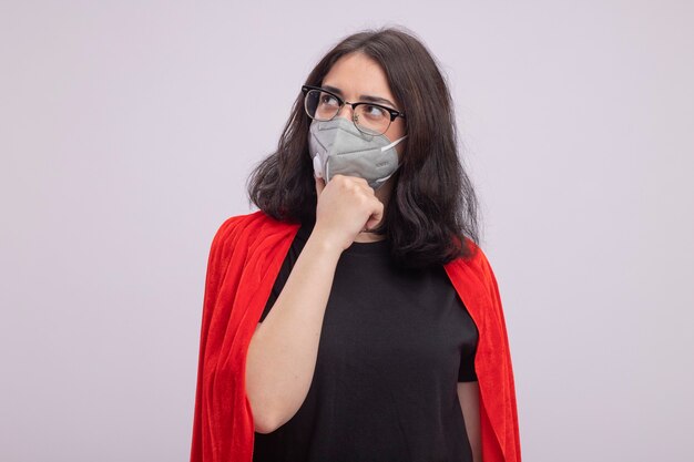 Thoughtful young caucasian superhero girl in red cape wearing glasses and protective mask keeping hand on chin looking at side isolated on white wall with copy space