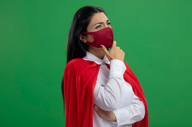 Thoughtful young caucasian superhero girl standing in profile view wearing mask putting hand on elbow and on chin looking at corner isolated on green background with copy space