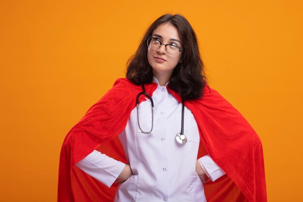 Free photo thoughtful young caucasian superhero girl wearing doctor uniform and stethoscope with glasses keeping hands on waist looking at side isolated on wall