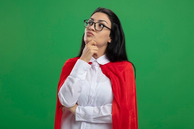 Thoughtful young caucasian superhero girl wearing glasses holding hand on elbow and on chin looking straigh isolated on green wall with copy space