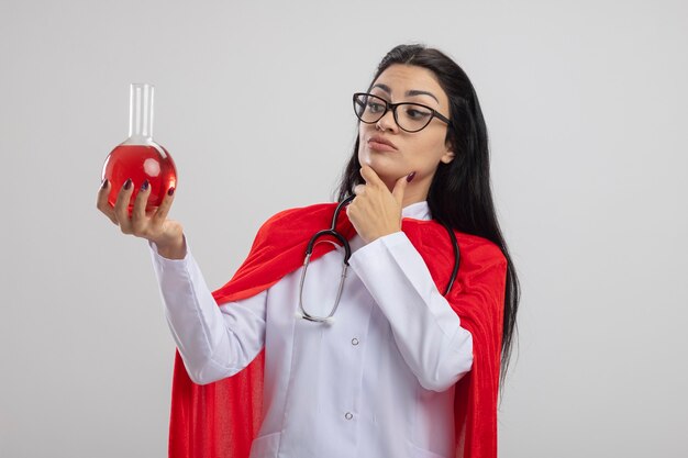 Thoughtful young caucasian superhero girl wearing glasses and stethoscope holding and looking at chemical flask with red liquid touching chin isolated on white background with copy space