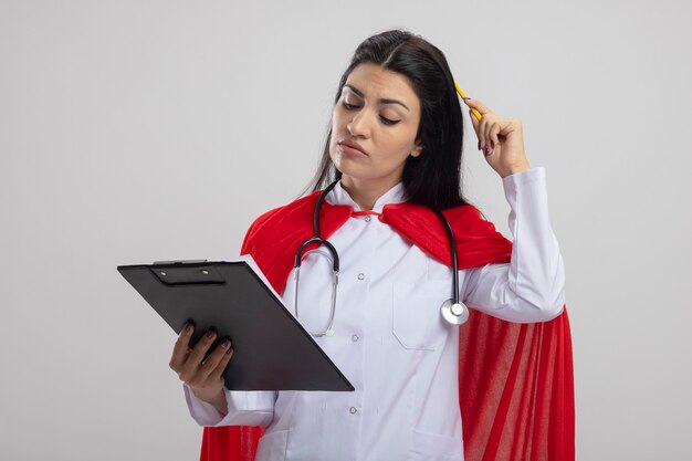 Thoughtful young caucasian superhero girl wearing stethoscope touching head with pencil holding and looking at clipboard isolated on white background