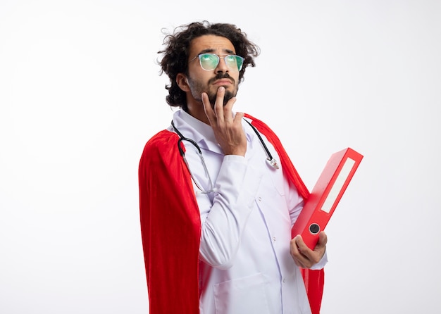 Thoughtful young caucasian superhero man in optical glasses wearing doctor uniform with red cloak and with stethoscope around neck puts hand on chin and holds file folder with copy space