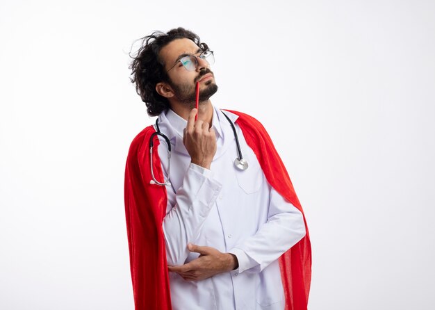 Thoughtful young caucasian superhero man in optical glasses wearing doctor uniform with red cloak and with stethoscope around neck puts pencil on his lip looking at side isolated on white wall