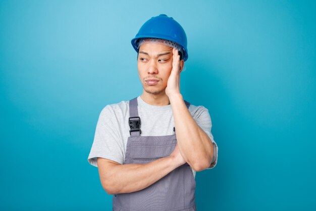 Thoughtful young construction worker wearing safety helmet and uniform keeping hand on head looking at side 