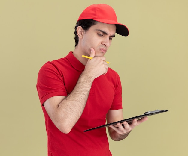 Thoughtful young delivery man in red uniform and cap holding clipboard and pencil looking at clipboard touching chin isolated on olive green wall