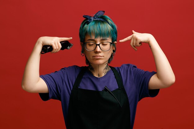 Thoughtful young female barber wearing uniform glasses and headband holding hair trimmer making brainstorming gesture with closed eyes isolated on red background