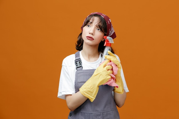 Free photo thoughtful young female cleaner wearing uniform rubber gloves and bandana holding cleanser and cloth duster looking at side touching face with cleanser isolated on orange background