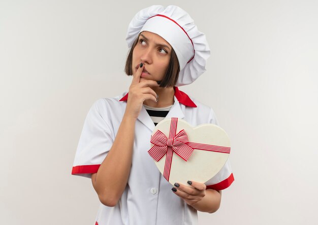 흰 벽에 고립 된 입술에 손가락으로 측면을보고 하트 모양의 선물 상자를 들고 요리사 유니폼 사려 깊은 젊은 여성 요리사