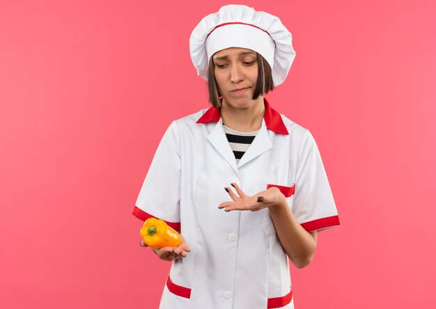 Задумчивая молодая женщина-повар в униформе шеф-повара смотрит и показывает рукой на перец, изолированный на розовой стене