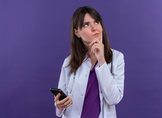 聴診器と医療ローブの思いやりのある若い女性医師は、電話を保持し、コピースペースで孤立した紫色の背景のあごに手を置きます