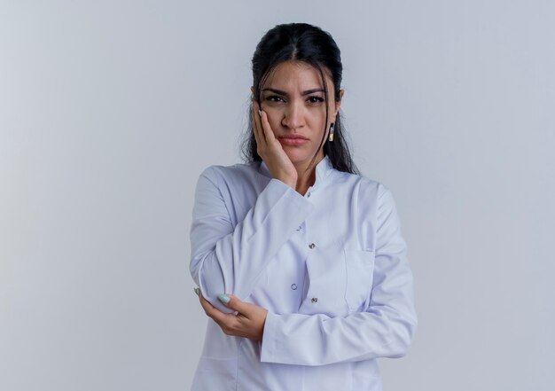 Thoughtful young female doctor wearing medical robe looking putting hand on elbow and keeping another one on face isolated