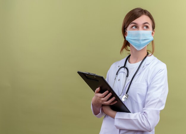 Задумчивая молодая женщина-врач в медицинском халате, маске и стетоскопе держит буфер обмена и смотрит на правую сторону на изолированной зеленой стене с копией пространства