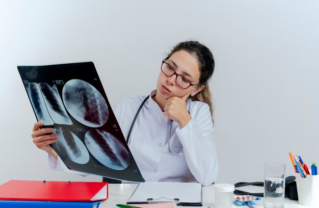 Thoughtful young female doctor wearing medical robe and stethoscope and glasses sitting at desk with medical tools holding looking at x-ray shot putting hand on chin isolated