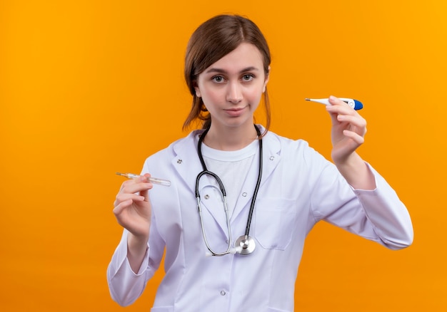 Thoughtful young female doctor wearing medical robe and stethoscope and holding thermometer with raised finger on isolated orange wall with copy space