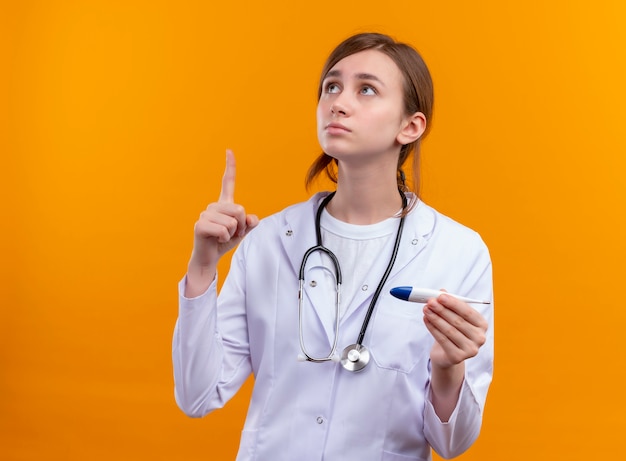 Giovane medico femminile premuroso che indossa veste medica e stetoscopio e che tiene il termometro con il dito alzato sulla parete arancione isolata con lo spazio della copia