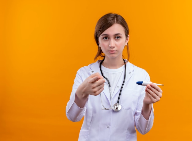 医療ローブと聴診器を身に着けて、コピースペースのある孤立したオレンジ色の壁に上げられた指で体温計を保持している思いやりのある若い女性医師