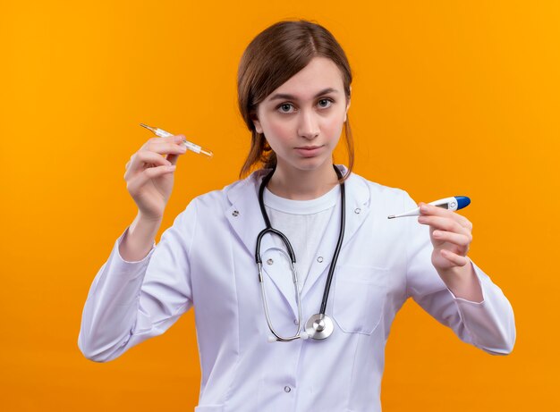 Задумчивая молодая женщина-врач в медицинском халате и стетоскопе, держащая термометр с поднятым пальцем на изолированной оранжевой стене с копией пространства