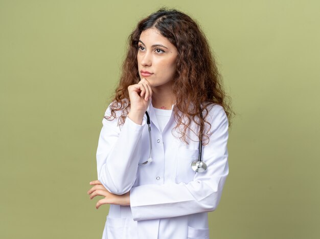 Задумчивая молодая женщина-врач в медицинском халате и стетоскопе держит руку на подбородке, глядя в сторону, изолированную на оливково-зеленой стене с копией пространства