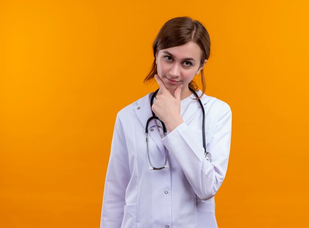 Thoughtful young female doctor wearing medical robe and stethoscope putting hand on chin on isolated orange wall with copy space