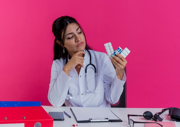 Thoughtful young female doctor wearing medical robe and stethoscope sitting at desk with medical tools holding and looking at medical drugs touching chin isolated on pink wall
