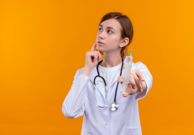 コピースペースのある孤立したオレンジ色の壁の左側を見て医療ローブと聴診器ストレッチ注射器を身に着けている思いやりのある若い女性医師