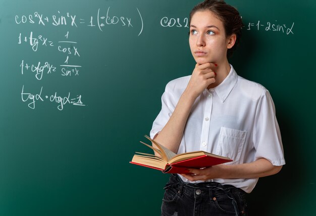 thoughtful young female math teacher standing in front of chalkboard holding open book touching chin looking at side in classroom